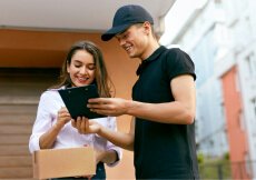 A delivery man in a cap shows a digital tablet to a smiling woman holding a parcel, both standing outdoors.