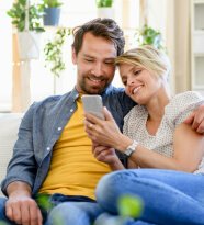 A couple sitting on a couch, smiling while looking at a smartphone together.