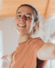 Young woman in a peach t-shirt smiling and extending her arm forward as if taking a selfie, with a softly blurred background.